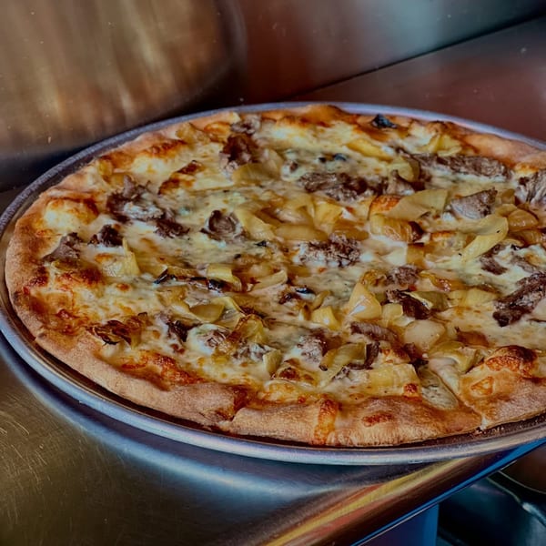 A freshly baked pizza with white sauce, cheese, steak, and caramelized onions on a metal tray.