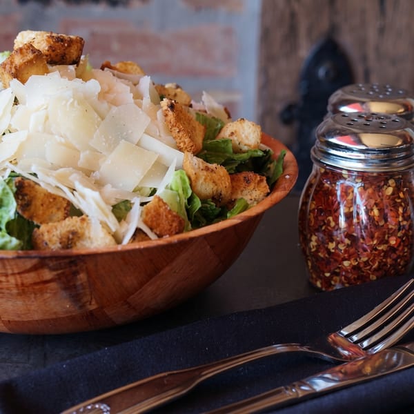 Caesar salad with shaved Parmesan and croutons in a wooden bowl, with red pepper flakes and cutlery.